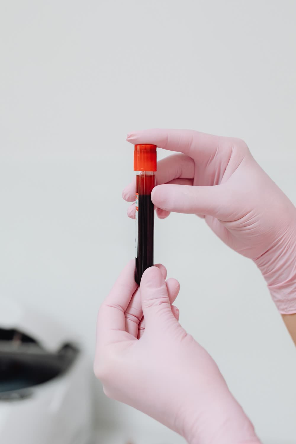 Gloved hands holding a blood collection tube with a red cap against a soft white background